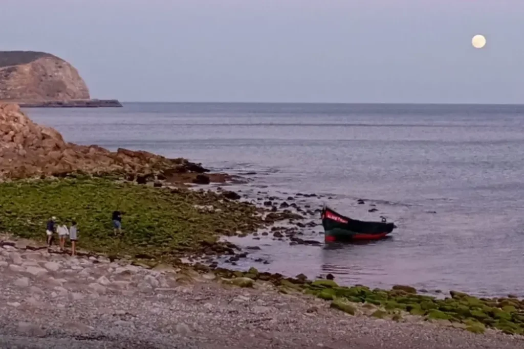 Il barcone arenato sulla spiaggia protagonista dello sbarco di migranti in Algarve, si vedono degli scogli e la luna nel cielo.