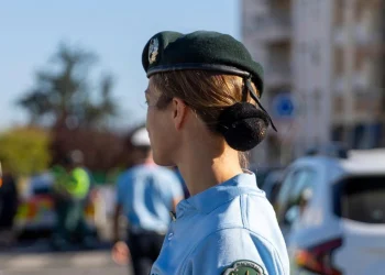 Su una strada un agente donna della GNR che indaga sulle truffe negli affitti, sullo sfondo alcune case vacanze e altri poliziotti.