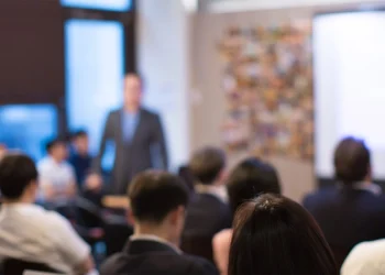 Un'immagine sfocata mostra un gruppo di persone sedute ad ascoltare un oratore nella parte anteriore di una stanza, forse durante una presentazione sull'accesso istruzione Portogallo. Sullo sfondo è visibile uno schermo luminoso.