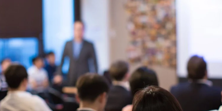 Un'immagine sfocata mostra un gruppo di persone sedute ad ascoltare un oratore nella parte anteriore di una stanza, forse durante una presentazione sull'accesso istruzione Portogallo. Sullo sfondo è visibile uno schermo luminoso.