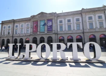 La grande insegna bianca "U. PORTO" si trova davanti a un grande edificio storico con finestre ad arco e striscioni colorati. In mezzo al brusio, gli studenti discutono della recente bufera facoltà medicina Porto sotto un cielo limpido e soleggiato.