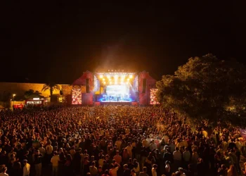 Una grande folla si riunisce in occasione di un concerto notturno all'aperto durante il Festival F Faro, di fronte a un palco illuminato con luci brillanti, circondato da alberi e strutture del festival.