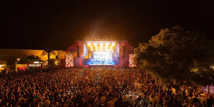Una grande folla si riunisce in occasione di un concerto notturno all'aperto durante il Festival F Faro, di fronte a un palco illuminato con luci brillanti, circondato da alberi e strutture del festival.