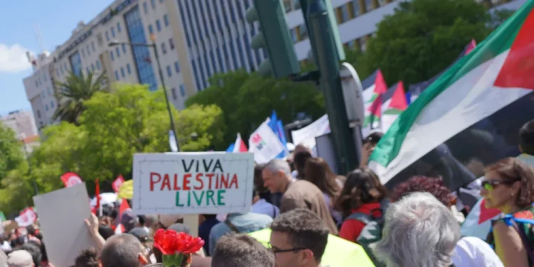 Una folla di manifestanti si riunisce all'aperto con cartelli e bandiere palestinesi, uno dei quali recita "Viva Palestina Livre" in portoghese, mentre Portogallo riconosce lo Stato Palestina. Un garofano rosso è attaccato al cartello. Sullo sfondo, edifici urbani.
