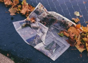 Un giornale accartocciato giace sul marciapiede in Portogallo, bagnato vicino a una rete di recinzione, circondato da foglie autunnali sparse: un sottile promemoria di come gli adulti analfabeti funzionali possano perdere questi momenti. La parola "SPORTS" è visibile.