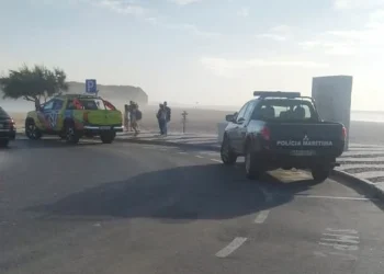 Su una strada due mezzi dei soccorsi, uno giallo e l'altro blu, sono parcheggiati per aiutare dei bagnini in pericolo. Si vedono alcune persone in attesa e il cielo è parzialmente nuvoloso.
