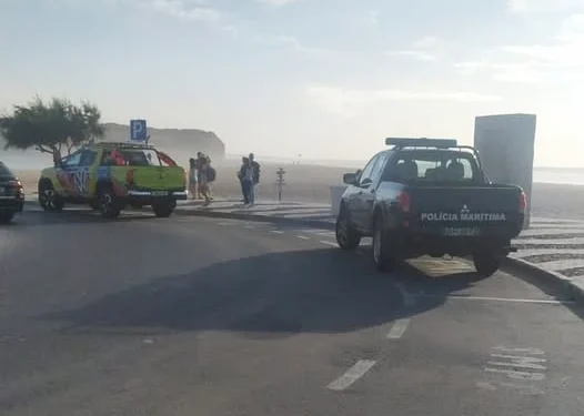 Su una strada due mezzi dei soccorsi, uno giallo e l'altro blu, sono parcheggiati per aiutare dei bagnini in pericolo. Si vedono alcune persone in attesa e il cielo è parzialmente nuvoloso.