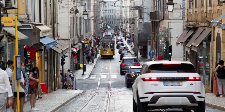 Una strada stretta e trafficata di Lisbona con automobili, un tram giallo, negozi su entrambi i lati e persone che camminano sui marciapiedi, soffrendo per la mancanza del controllo qualità dell'aria. Vecchi edifici fiancheggiano la strada mentre un SUV bianco passa, mentre il controllo qualità aria garantisce ambienti urbani più puliti.
