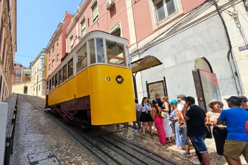 Un immagine della funicolare da Gloria di Lisbona deragliata, si vede la cabina ferma sui binari in mezzo a palazzi alti e alcune persone in attesa di salire.