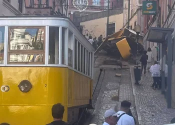 La funicolare di Lisbona e diverse persone si trovano in fondo a una strada stretta, dove una funicolare deragliata è precipitata e si è ribaltata davanti a noi, bloccando i binari. I detriti sono sparsi mentre gli astanti osservano il malfunzionamento e attendono la manutenzione.