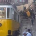 La funicolare di Lisbona e diverse persone si trovano in fondo a una strada stretta, dove una funicolare deragliata è precipitata e si è ribaltata davanti a noi, bloccando i binari. I detriti sono sparsi mentre gli astanti osservano il malfunzionamento e attendono la manutenzione.