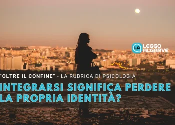 Una sagoma di donna in piedi su una collina che domina la città di Lisbona al tramonto, con testo italiano su psicologia, identità e integrazione dopo l' espatrio da "Leggo Algarve.