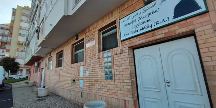 Esterno di un edificio in mattoni della moschea di Lisbona oggetto di minaccia, "Masjid (Moschea) Sayyidina Abu Bakr Siddiq (R.A.)" in inglese e arabo, con doppie porte bianche, finestre e marciapiede.