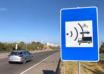 Un cartello che segnala un autovelox su una strada in Portogallo e una auto di color grigio che frena per non prendere una multa. Il cielo è azzurro e si vede della vegetazione sullo sfondo.