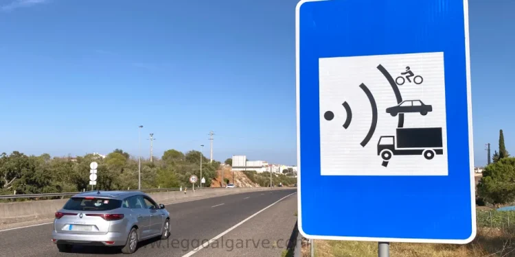 Un cartello che segnala un autovelox su una strada in Portogallo e una auto di color grigio che frena per non prendere una multa. Il cielo è azzurro e si vede della vegetazione sullo sfondo.
