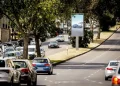 Un'automobile percorre una strada urbana alberata di Lisbona con auto parcheggiate su entrambi i lati. Un grande cartellone stradale, parte dei nuovi pannelli pubblicitari di Lisbona, pubblicizza un'automobile mentre la luce del sole filtra attraverso gli alberi sulla strada.