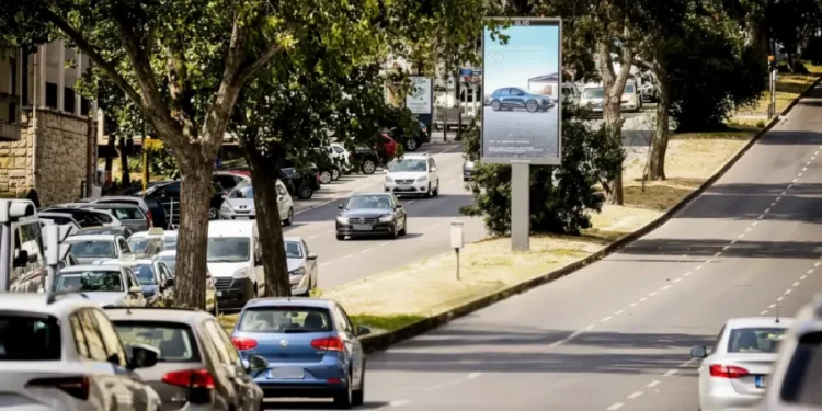 Un'automobile percorre una strada urbana alberata di Lisbona con auto parcheggiate su entrambi i lati. Un grande cartellone stradale, parte dei nuovi pannelli pubblicitari di Lisbona, pubblicizza un'automobile mentre la luce del sole filtra attraverso gli alberi sulla strada.