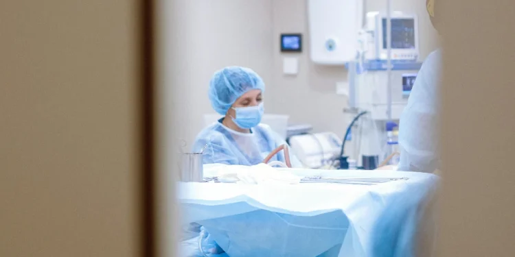 Un operatore sanitario in camice chirurgico, mascherina e copricapo è visto attraverso una porta del nuovo ospedale Lusiadas Faro, mentre prepara strumenti medici accanto a un paziente in una stanza di ospedale o clinica. Sullo sfondo sono visibili le attrezzature mediche.