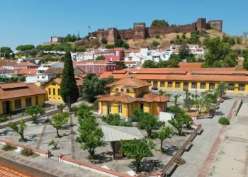 Veduta aerea di una città con edifici dai tetti arancioni, un cortile alberato e un grande castello medievale su una collina, circondato dal verde e da case bianche, che mostra l'impatto della riqualificazione di Fábrica do Inglês nella zona.