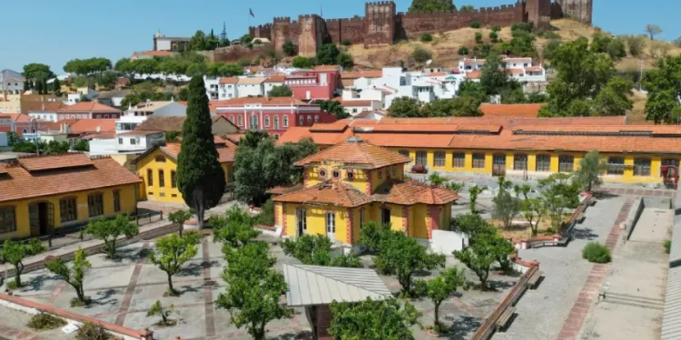 Veduta aerea di una città con edifici dai tetti arancioni, un cortile alberato e un grande castello medievale su una collina, circondato dal verde e da case bianche, che mostra l'impatto della riqualificazione di Fábrica do Inglês nella zona.