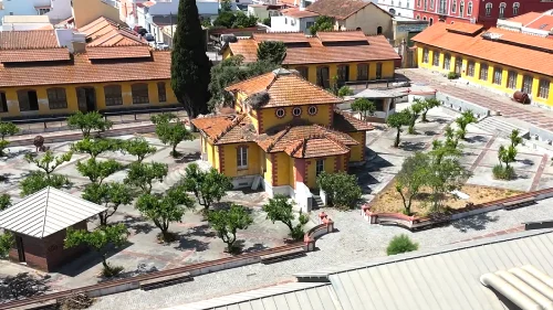 Veduta aerea di una città con edifici dai tetti arancioni, un cortile alberato e un grande castello medievale su una collina, circondato dal verde e da case bianche, che mostra l'impatto della riqualificazione di Fábrica do Inglês nella zona.