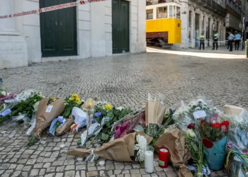 Mazzi di fiori, candele e biglietti vengono deposti su una strada acciottolata come memoriale dopo la tragedia di Lisbona, evidenziando l'impatto psicologico sulla comunità. Agenti di polizia sullo sfondo vicino a un tram giallo e a un edificio transennato.