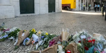 Mazzi di fiori, candele e biglietti vengono deposti su una strada acciottolata come memoriale dopo la tragedia di Lisbona, evidenziando l'impatto psicologico sulla comunità. Agenti di polizia sullo sfondo vicino a un tram giallo e a un edificio transennato.