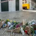 Mazzi di fiori, candele e biglietti vengono deposti su una strada acciottolata come memoriale dopo la tragedia di Lisbona, evidenziando l'impatto psicologico sulla comunità. Agenti di polizia sullo sfondo vicino a un tram giallo e a un edificio transennato.