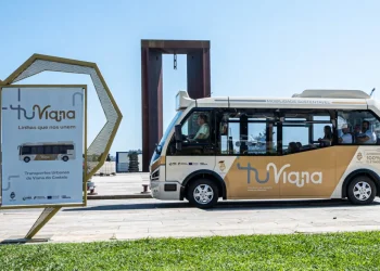 Un piccolo autobus del trasporto pubblico di Viana do Castelo, di colore beige e bianco con la scritta "tuViana", è parcheggiato vicino a una grande scultura e a un cartello promozionale con il marchio corrispondente, in una giornata di sole sul lungomare.