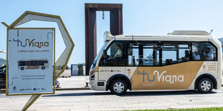 Un piccolo autobus del trasporto pubblico di Viana do Castelo, di colore beige e bianco con la scritta "tuViana", è parcheggiato vicino a una grande scultura e a un cartello promozionale con il marchio corrispondente, in una giornata di sole sul lungomare.