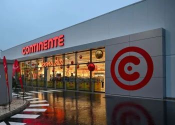 L'esterno di un supermercato Continente con la grande insegna rossa, le vetrine che espongono i prodotti all'interno e il logo rosso in grassetto sul muro grigio. La pavimentazione bagnata riflette l'edificio, le luci e accenna a preoccupazioni come la truffa con carta Continente.