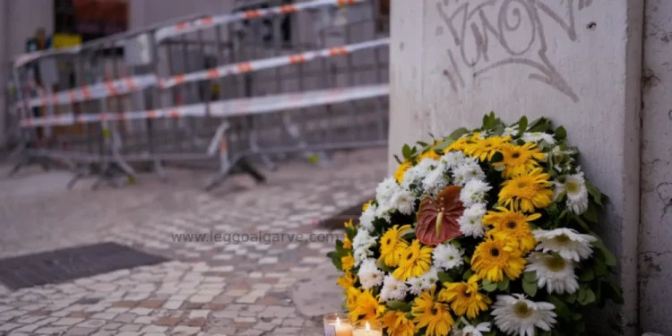 Un bouquet di fiori gialli e bianchi più due candele accese sono poste all'angolo della strada in memoria delle vittime della funicolare di Lisbona, si vedono sullo sfondo le transenne e i fettuccine della polizia.