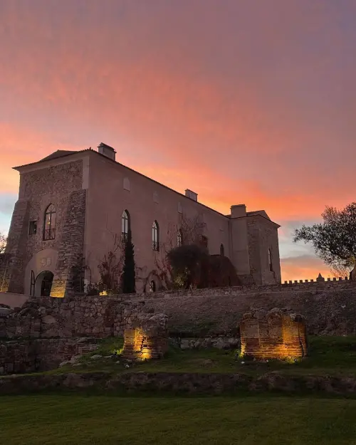 Un edificio storico in pietra si erge su una collina erbosa al tramonto, con le pareti immerse in una luce calda. Il cielo si tinge di rosa e arancio mentre i riflettori illuminano le rovine dell'Azienda Vinicola Fitapreta Évora in primo piano.