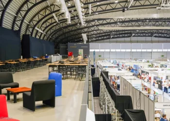 Immagine all'interno del Blip Expo Algarve, nella Portimão Arena, con un alto soffitto ad arco, stand espositivi sottostanti e un mezzanino con tavoli, sedie e poltrone colorate. Le persone passeggiano e visitano gli stand al livello inferiore.