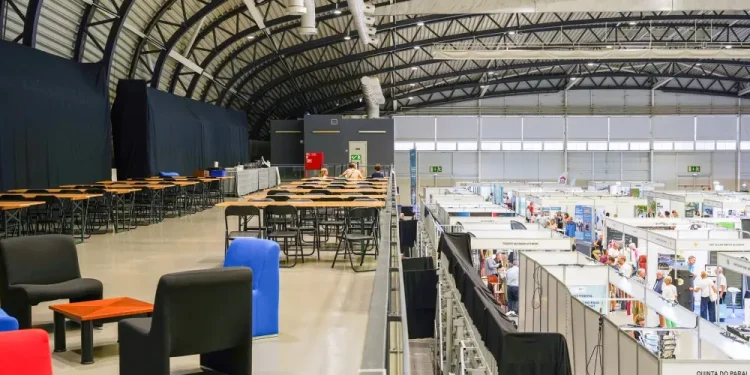 Immagine all'interno del Blip Expo Algarve, nella Portimão Arena, con un alto soffitto ad arco, stand espositivi sottostanti e un mezzanino con tavoli, sedie e poltrone colorate. Le persone passeggiano e visitano gli stand al livello inferiore.