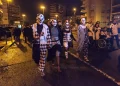 Un gruppo di persone vestite con costumi da clown bianchi e neri e dipinti sul viso camminano insieme di notte in una strada di Lisbona in Portgallo durante la festa di Halloween, circondati da altre persone e da alti edifici.
