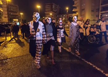 Un gruppo di persone vestite con costumi da clown bianchi e neri e dipinti sul viso camminano insieme di notte in una strada di Lisbona in Portgallo durante la festa di Halloween, circondati da altre persone e da alti edifici.