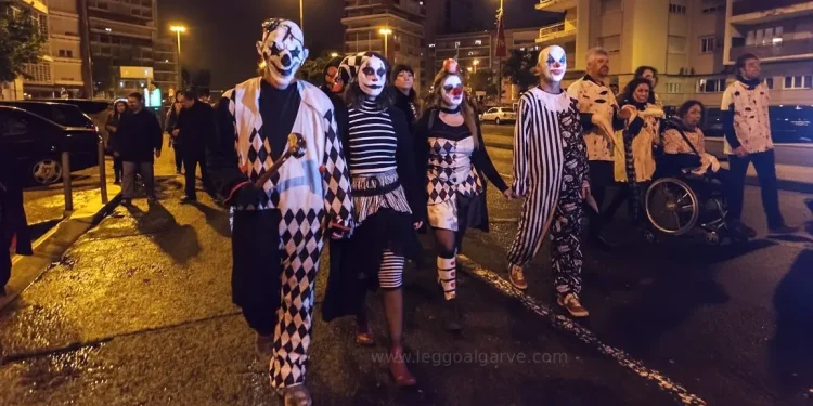 Un gruppo di persone vestite con costumi da clown bianchi e neri e dipinti sul viso camminano insieme di notte in una strada di Lisbona in Portgallo durante la festa di Halloween, circondati da altre persone e da alti edifici.