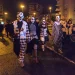 Un gruppo di persone vestite con costumi da clown bianchi e neri e dipinti sul viso camminano insieme di notte in una strada di Lisbona in Portgallo durante la festa di Halloween, circondati da altre persone e da alti edifici.