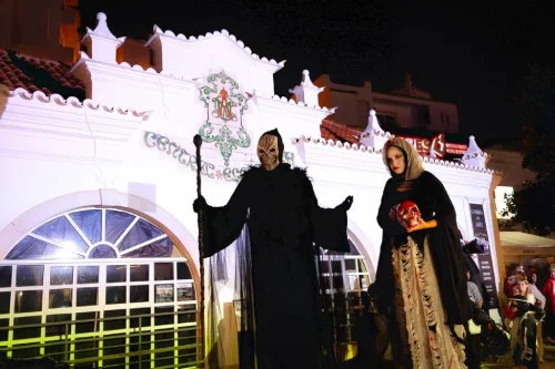 Due persone con elaborati costumi di Halloween, uno come un tristo mietitore e l'altro come una figura gotica con un teschio rosso, si trovano di fronte a un edificio bianco di notte, illuminato a giorno, catturando l'inquietante spirito di Halloween in Portogallo.