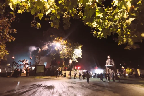 Una scena notturna all'aperto a Vilar de Perdizes mostra un gruppo di persone riunite vicino a un edificio illuminato e a una grande statua; rami frondosi incorniciano la parte superiore dell'immagine, evocando la festa di Halloween in Portogallo.