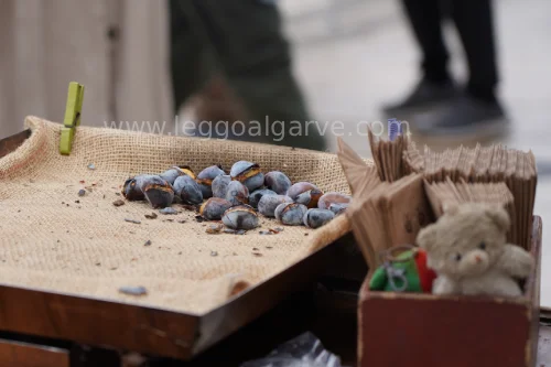 Un ripiano rivestito di iuta con alcune castagne arrosto che nel periodo di Halloween in Portogallo sono molto comuni. Si vede sfuocato sullo sfondo una strada con i piedi di una persona e un piccolo peluche in una cassettina di legno. 
