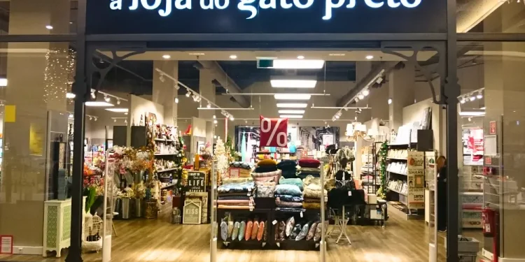 La vetrina di Loja Gato Preto presenta un interno luminoso. All'ingresso, cuscini, fiori e oggetti decorativi sono disposti con cura, mentre un cartello di vendita rosso con un simbolo di percentuale evidenzia la situazione degli sconti in corso.