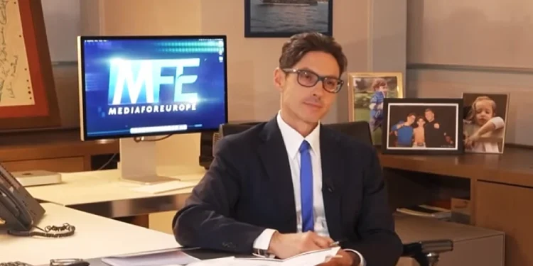 Piersilvio Berlusconi in giacca e cravatta e occhiali è seduto a una scrivania con documenti, telefono e materiale da ufficio, mentre si discute di Mfe che vorrebbe entrare nel mercato portoghese. Dietro di lui c'è il monitor di un computer con la scritta "MFE MEDIAFOREUROPE", foto di famiglia sulla parete.