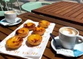 Due tazze di caffè e sei crostate portoghesi (pastéis de nata) di un punto Miglior Pastel Nata Lisbona sono disposte su un tavolo di legno all'aperto di un bar, con sedie e una strada lastricata sullo sfondo.