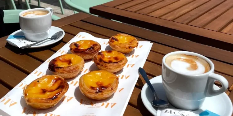 Due tazze di caffè e sei crostate portoghesi (pastéis de nata) di un punto Miglior Pastel Nata Lisbona sono disposte su un tavolo di legno all'aperto di un bar, con sedie e una strada lastricata sullo sfondo.