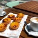 Due tazze di caffè e sei crostate portoghesi (pastéis de nata) di un punto Miglior Pastel Nata Lisbona sono disposte su un tavolo di legno all'aperto di un bar, con sedie e una strada lastricata sullo sfondo.
