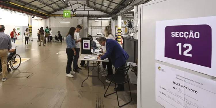 Persone che votano in un seggio elettorale di Portogallo durante la domenica vota amministrativa, con il cartello "Seção 12" (Sezione 12) visibile ed elettori e funzionari riuniti in un'ampia area interna.