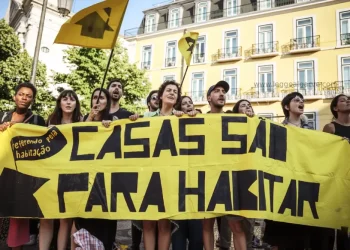 Un gruppo di persone protesta all'aperto contro i prezzi esagerati di Portogallo, tenendo in mano un grande striscione giallo con la scritta "CASAS SÃO PARA HABITAR" e bandiere gialle. Sullo sfondo appare un edificio con balconi.