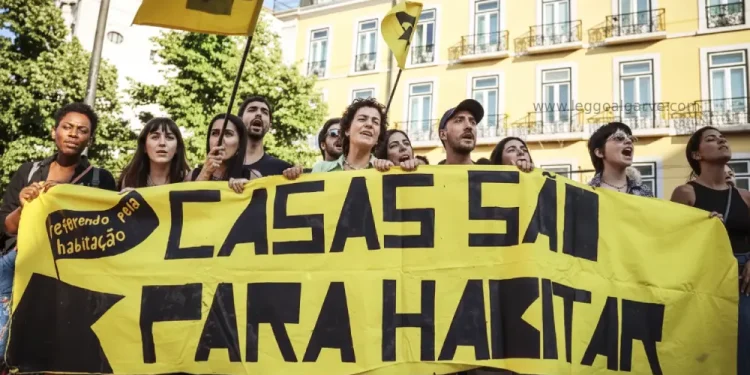 Un gruppo di persone protesta all'aperto contro i prezzi esagerati di Portogallo, tenendo in mano un grande striscione giallo con la scritta "CASAS SÃO PARA HABITAR" e bandiere gialle. Sullo sfondo appare un edificio con balconi.